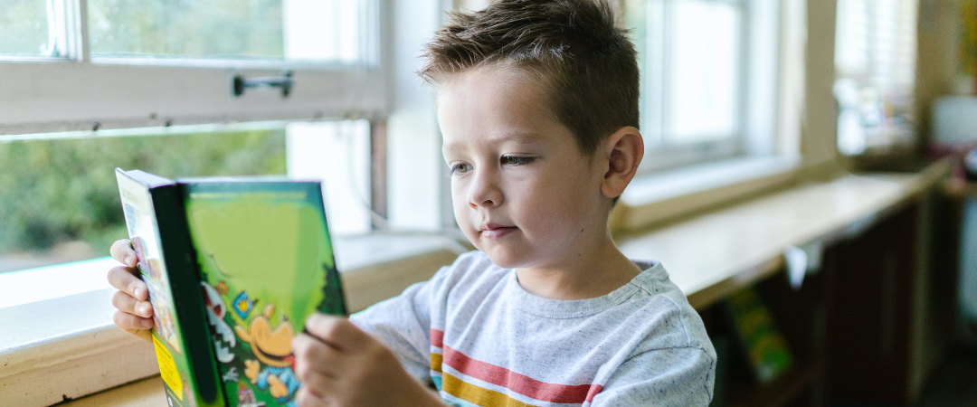 A young boy engrossed in reading a book in a classroom, surrounded by desks and educational materials.