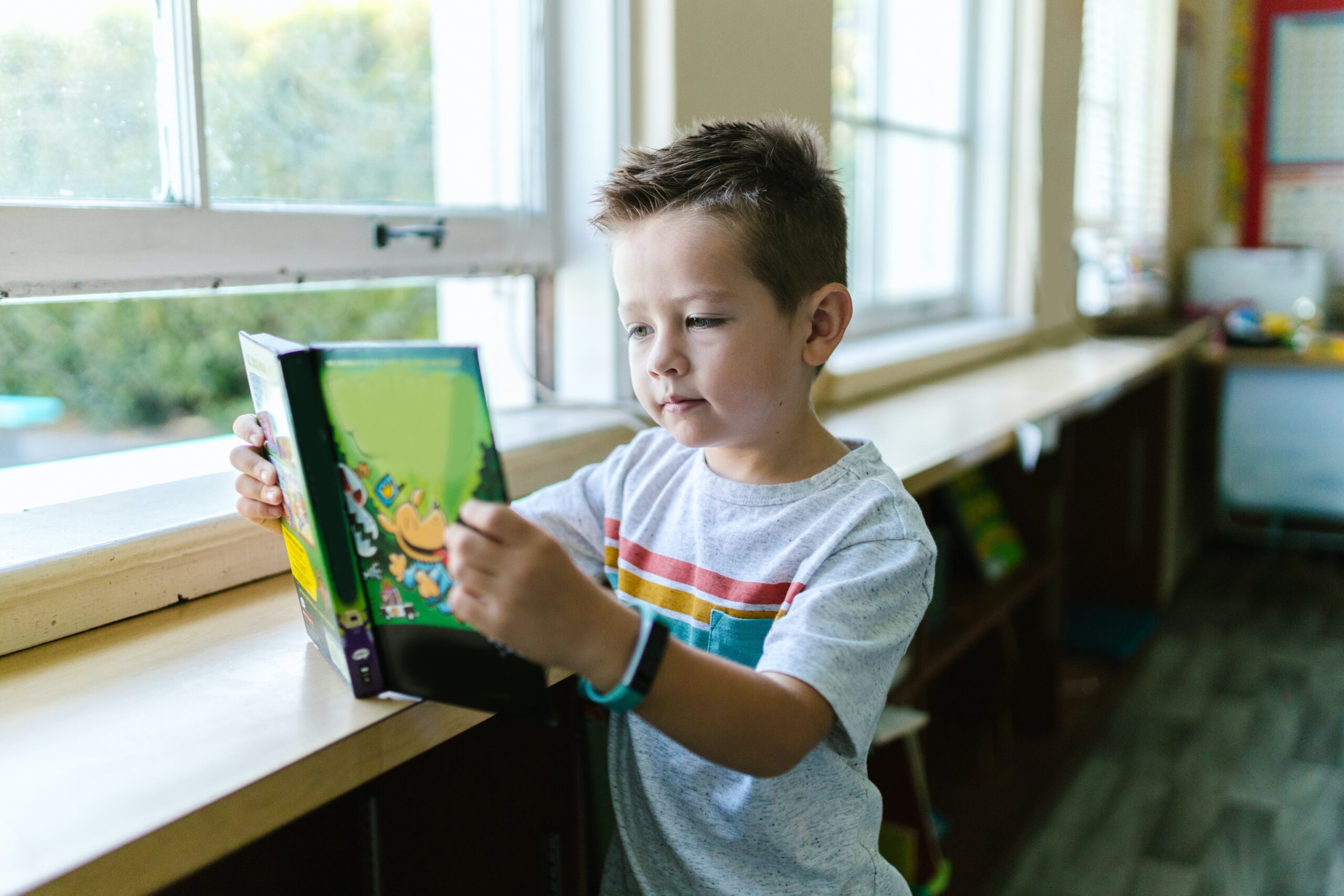 
A young boy engrossed in reading a book in a classroom, surrounded by desks and educational materials.